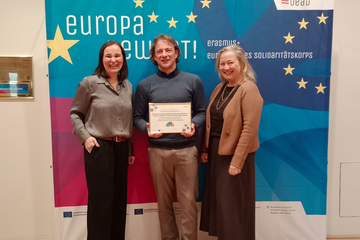 Zwei Frauen und ein Mann sthen vor einenm Plakat 