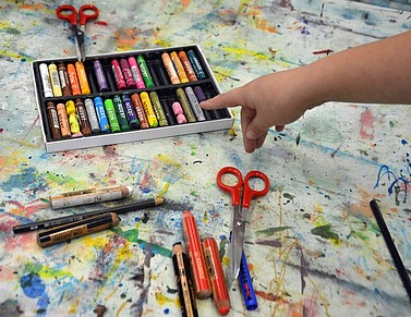 Offener Farbmalkasten mit Stiften auf Maldecke mit Farbspritzern,, lose Stifte in verschiedenen Farben, 2 Scheren, Hand zeigt auf Stifte