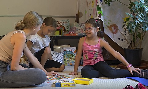 2 Mädchen sitzen mit Frau am Boden und spielen Spiel, im Hintergrund andere Spiele 