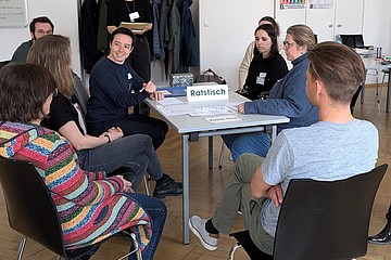 Pädagog*innen sitzen um einen Tisch, auf dem ein Aufsteller mit "Ratstisch" steht. 