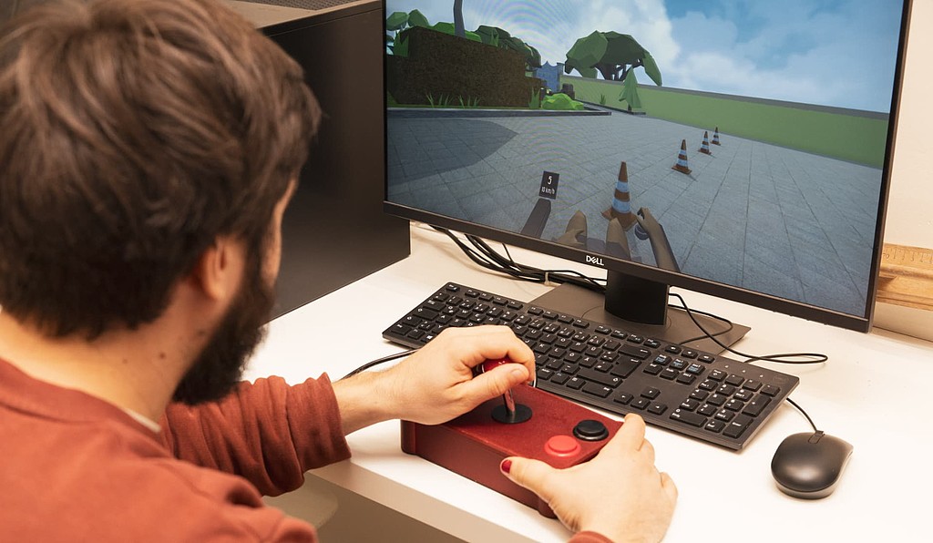 Ein Studierender nutzt ein assistives Joystick-Eingabegerät, um eine virtuelle Simulation auf dem Monitor zu steuern. Das Bild verdeutlicht die praktische Erprobung von Hilfsmitteln für Menschen mit motorischen Einschränkungen im Rahmen der Hochschullernwerkstatt.