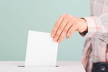 Hand steckt Wahlkuvert in Wahlurne